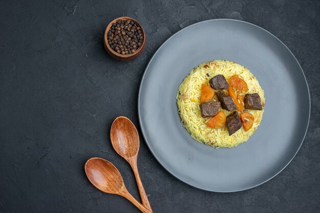 Vista de cima, delicioso pilaf cozido de arroz com damascos secos e fatias de carne dentro do prato em superfície escura