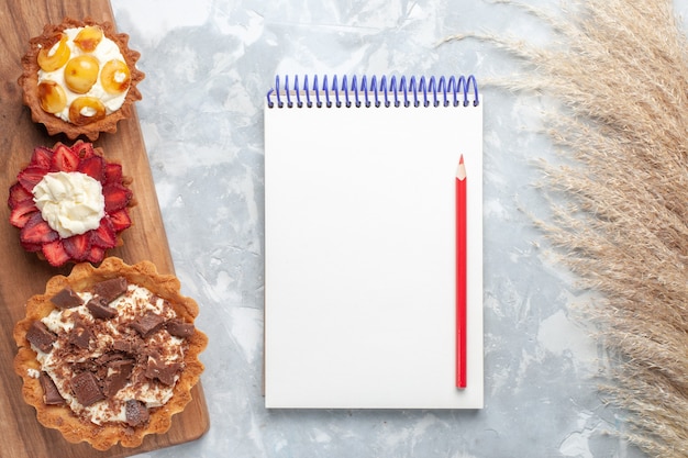 Vista de cima diferentes bolos cremosos com bloco de notas de chocolate e frutas na mesa branca bolo assar biscoito doce açúcar fruta