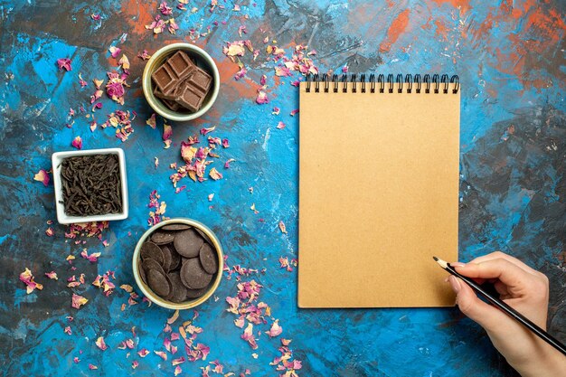 Vista de cima diferentes chocolates em pequenas tigelas, lápis de caderno na mão da mulher na superfície azul vermelha