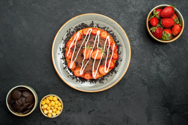 Foto grátis vista de cima do bolo com placa de chocolate do bolo com chocolate e morango entre tigelas de avelã de morango e chocolate na mesa escura