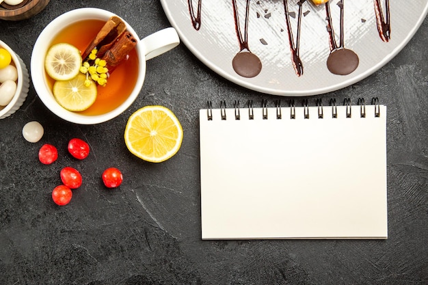 Foto grátis vista de cima doces e chá xícara de chá branco com paus de canela e limão ao lado do caderno branco o prato de bolo e tigelas de chocolate e doces na mesa escura