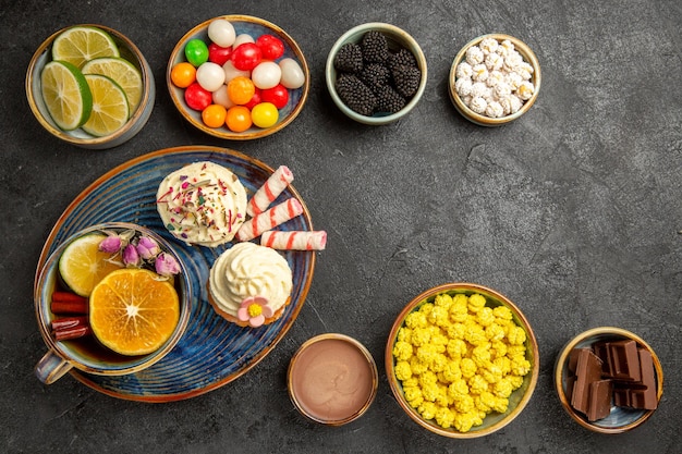 Vista de cima doces na mesa tigelas de frutas cítricas, chocolate, doces coloridos e um prato de cupcakes e uma xícara de chá de ervas com paus de canela na mesa escura