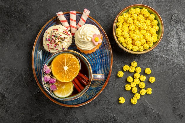 Vista de cima doces no prato cupcakes com creme branco no pires uma xícara de chá com limão e canela em pau tigela de doces amarelos na mesa