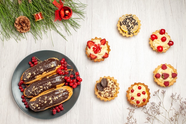 Vista de cima, éclairs de chocolate e groselhas nas tortas de prato cinza e folhas de pinheiro com brinquedos de natal no chão de madeira branco