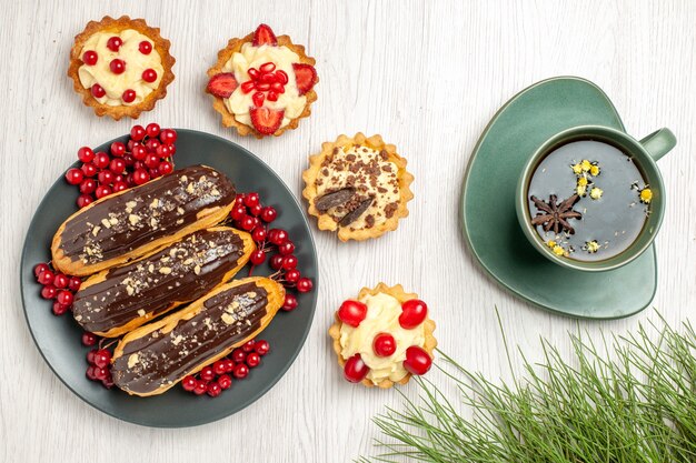 Vista de cima, éclairs de chocolate e groselhas no prato cinza cercado com tortas, uma xícara de chá e folhas de pinheiro na mesa de madeira branca