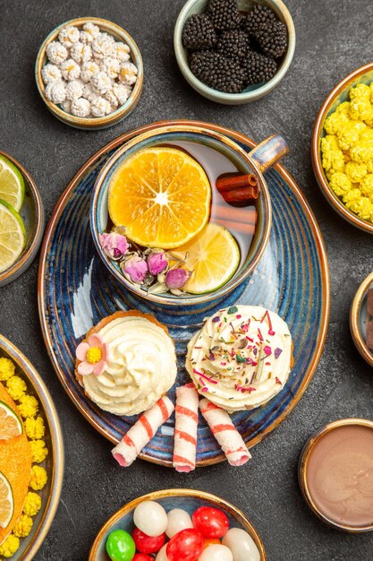 Vista de cima em close-up uma xícara de chá de ervas um prato de cupcakes com creme uma xícara de chá de ervas com limão e doces ao lado das tigelas de frutas cítricas doces brancos na mesa escura