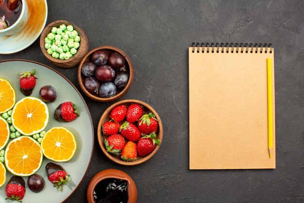 Vista de cima em close-up xícara de chá e frutas uma xícara de chá com cobertura de chocolate, morango picado de laranja e balas verdes e tigelas com diferentes frutas e doces ao lado do caderno e lápis