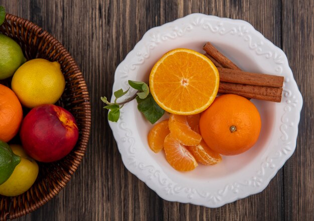 Vista de cima fatias de laranja descascadas com canela em um prato e limão e pêssego em uma cesta com fundo de madeira