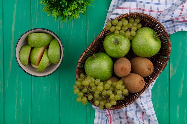 Vista de cima fatias de maçã verde em uma tigela com kiwi e uvas em cestas sobre fundo verde