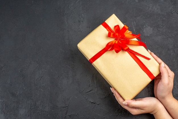 Vista de cima, mãos femininas segurando um presente de Natal em papel pardo amarrado com fita vermelha na superfície escura