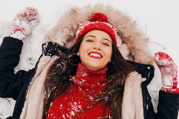 Vista de cima na jovem mulher feliz e sorridente, muito sincera, com luvas vermelhas e chapéu de malha, vestindo um casaco preto deitado na neve no parque, roupas quentes, se divertindo