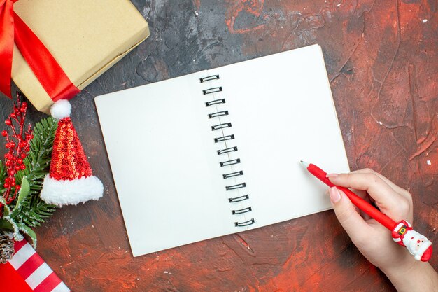 Vista de cima para presentes de Natal amarrados com fita vermelha e caneta de bloco de notas de chapéu de Papai Noel na mão feminina na mesa vermelha escura