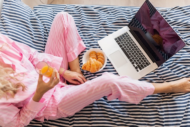 Vista de cima para uma jovem loira e bonita de pijama rosa sentada na cama com laptop tomando café