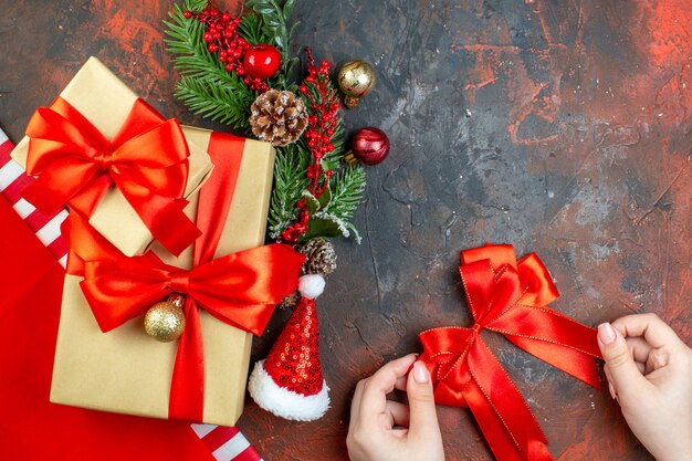 Vista de cima pequenos presentes amarrados com fita vermelha chapéu de Papai Noel galho de árvore de natal laço vermelho na mão feminina na mesa vermelho escuro