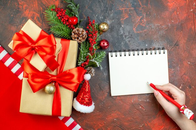 Vista de cima pequenos presentes amarrados com fita vermelha, chapéu de Papai Noel, ramo de árvore de Natal, bloco de notas caneta vermelha na mesa vermelha escura