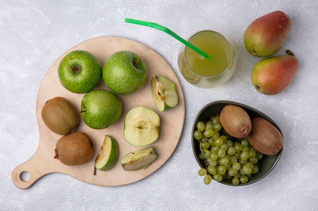 Vista de cima peras com kiwi e uvas em uma tigela com fatias de maçã verde em um suporte com suco de maçã em um fundo branco