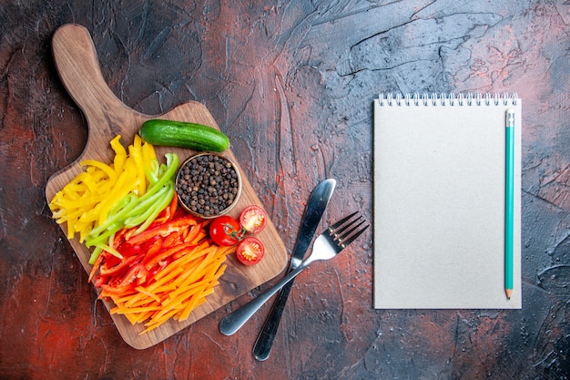 Vista de cima pimentão colorido cortado pimenta preta tomate pepino na tábua de cortar lápis no garfo do caderno e faca na mesa vermelho escuro