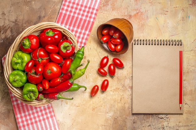 Vista de cima pimentas verdes e vermelhas, pimentas, tomates em uma cesta de vime, tomates cereja espalhados da tigela, toalha de cozinha, um caderno e lápis vermelho sobre fundo âmbar