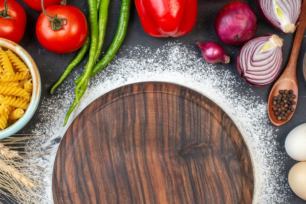 Vista de cima placa de madeira polvilhada com massa espiral de farinha na tigela ovos cebola vermelha pimenta preta em colher de pau tomates na mesa