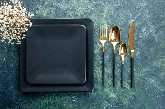 vista de cima pratos quadrados pretos com garfo dourado colheres e faca em fundo azul escuro jantar cozinha restaurante comida talheres cor