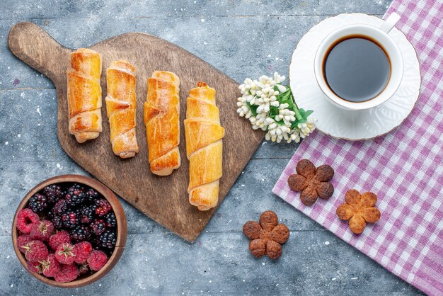 Vista de cima, pulseiras deliciosas e doces com recheio, juntamente com uma xícara de café e biscoitos, bagas na mesa de madeira cinza.