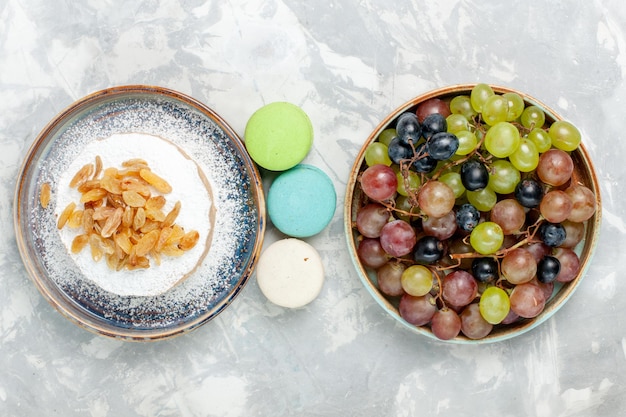 Foto grátis vista de cima redondo pequeno bolo de açúcar em pó com passas, macarons franceses e uvas na mesa branca