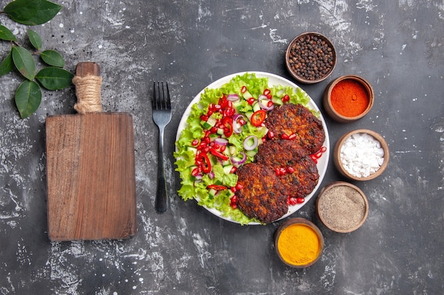 Vista de cima saborosas costeletas de carne com salada e temperos em fundo cinza foto comida prato de carne