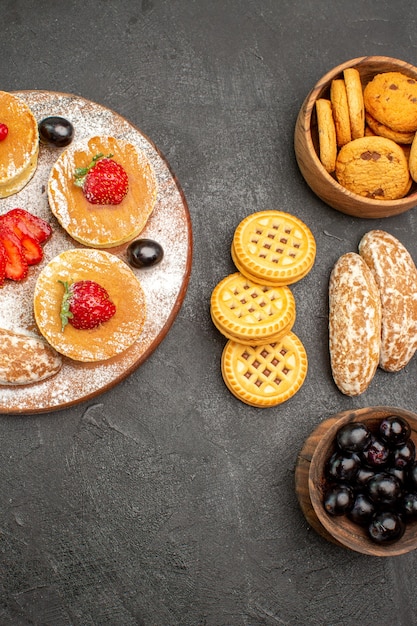 Vista de cima saborosas panquecas com bolos doces e frutas em uma superfície escura, sobremesa de bolos de açúcar