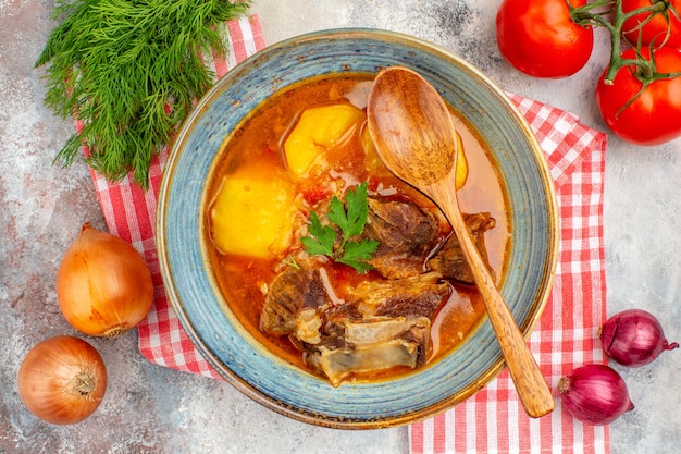 Vista de cima sopa bozbash caseira, colher de pau, toalha de cozinha, um monte de cebolas de tomate endro em fundo nu