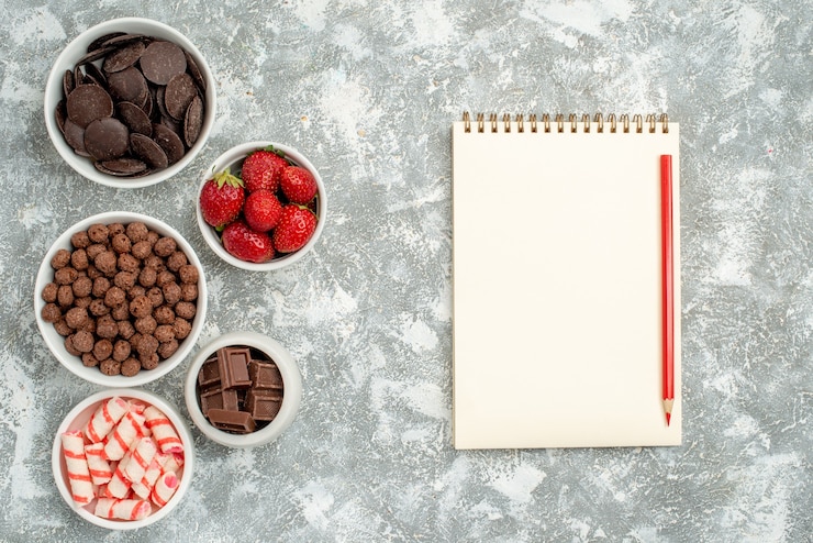 Foto grátis vista de cima tigelas com doces, morangos, chocolates, cereais e cacau à esquerda e um caderno com lápis vermelho à direita da mesa cinza-esbranquiçada