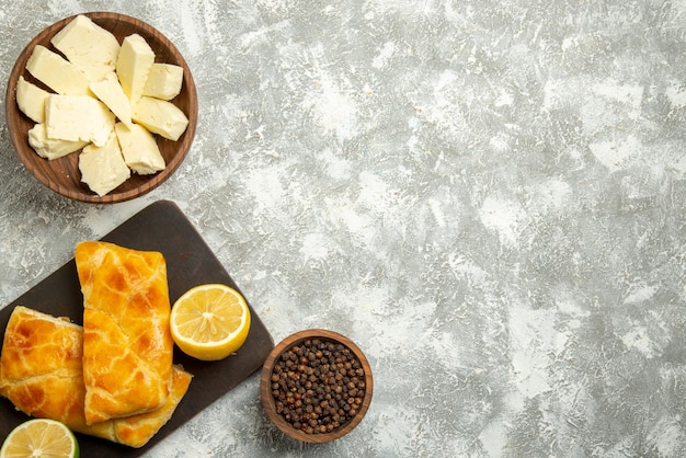 Vista de cima tortas de queijo tigelas de madeira com queijo e pimenta preta, tortas apetitosas e limão na tábua de corte no lado esquerdo da mesa