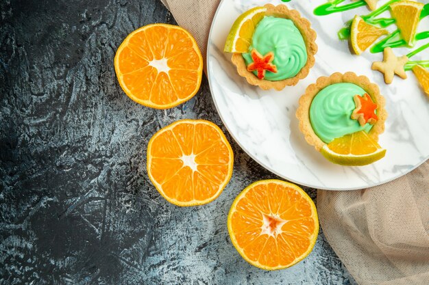 Vista de cima tortinhas com creme verde e fatia de limão no prato sobre xale bege, corte laranjas na mesa escura.