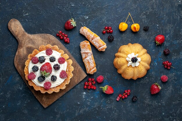 Vista de cima um bolo gostoso com creme e frutas vermelhas junto com pulseiras na mesa escura.