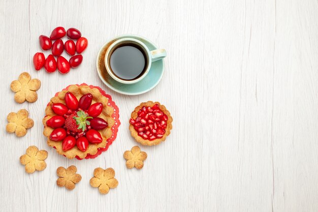 Vista de cima um pequeno bolo com biscoitos de frutas e uma xícara de chá na mesa branca