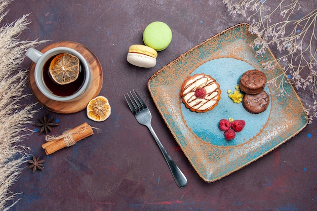 Vista de cima um pequeno bolo cremoso com biscoitos de chocolate e uma xícara de chá em uma superfície escura.