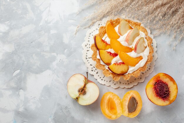 Vista de cima um pequeno bolo cremoso com frutas fatiadas e creme branco, juntamente com damascos frescos e pêssegos em um biscoito de mesa com luz branca