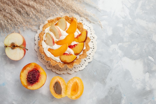Vista de cima um pequeno bolo cremoso com frutas fatiadas e creme branco junto com damascos frescos e pêssegos em biscoito de frutas de mesa com luz branca