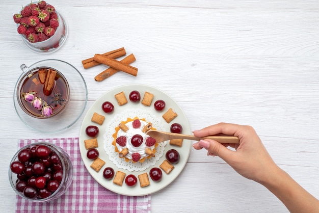 Vista de cima um pequeno bolo delicioso com framboesas, cerejas e bolachas chá de canela na mesa branca frutas baga creme chá