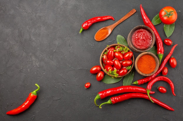 Vista de cima uma tigela de tomates cereja pimentas vermelhas uma colher de pau folhas de louro tigelas de ketchup e pimenta em pó e tomate no chão preto