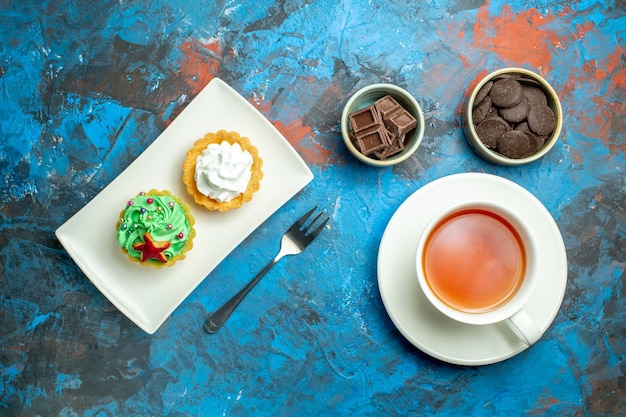 Vista de cima uma xícara de bolos de chá em um prato de chocolates em pequenas tigelas na superfície vermelha azul