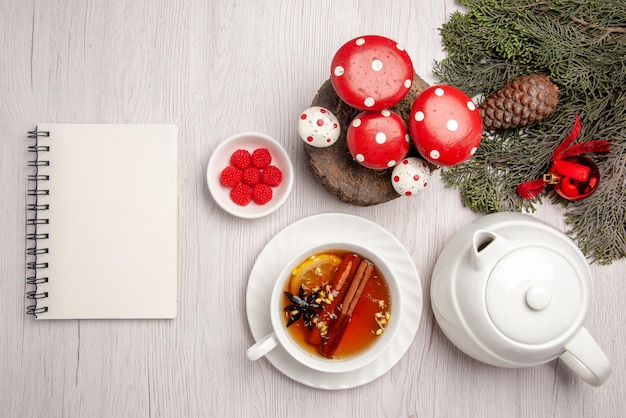 Vista de cima uma xícara de chá de chá de ervas com limão e canela na xícara ao lado do caderno branco de bagas de bule e galhos de árvore de natal com cone e brinquedos de árvore de natal em cima da mesa