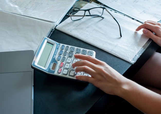 Vista de close-up de mãos femininas fazendo cálculos e documentos com óculos na mesa de escritório, processo de trabalho, conceito de escritório