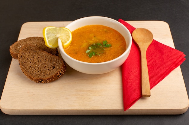 Vista de frente saborosa sopa de vegetais dentro do prato com uma fatia de limão e pães na mesa escura.