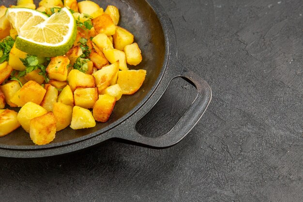 Vista de frente saborosas batatas fritas dentro de uma panela com rodelas de limão na mesa escura