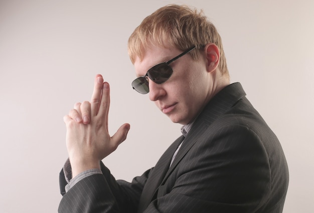 Vista de um homem vestindo um terno preto e óculos escuros enquanto faz a posição de uma arma com os dedos