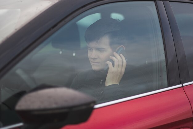 Vista de um jovem homem vestindo uma jaqueta preta sentado em um carro vermelho enquanto fala ao telefone