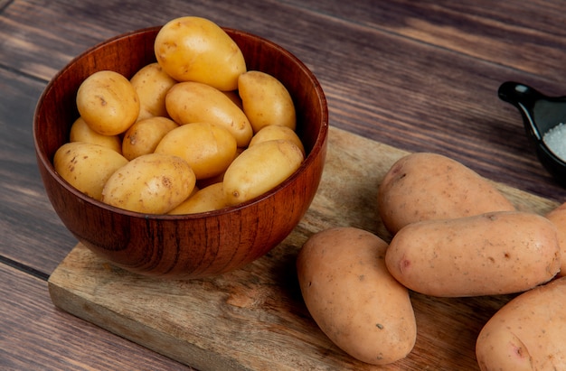 Vista do close-up de batatas novas na tigela e as brancas na tábua com sal na superfície de madeira