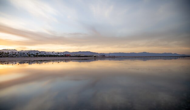 Vista do mar com um céu refletivo. O sol se põe sobre a cidade perto das montanhas.