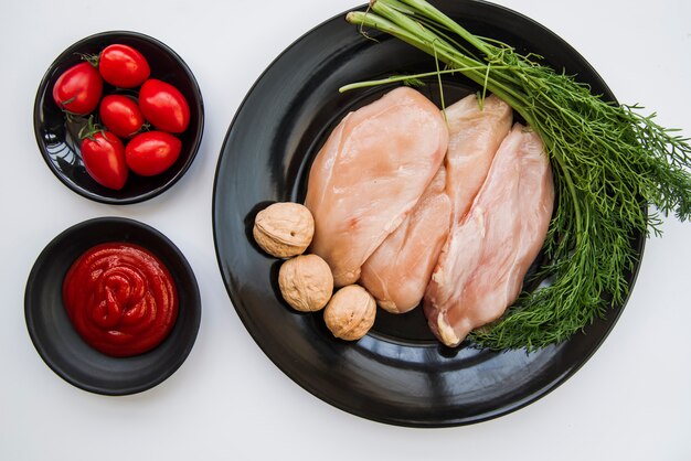 Vista elevada de frango cru; tempero fresco; noz; tomate e molho no fundo branco