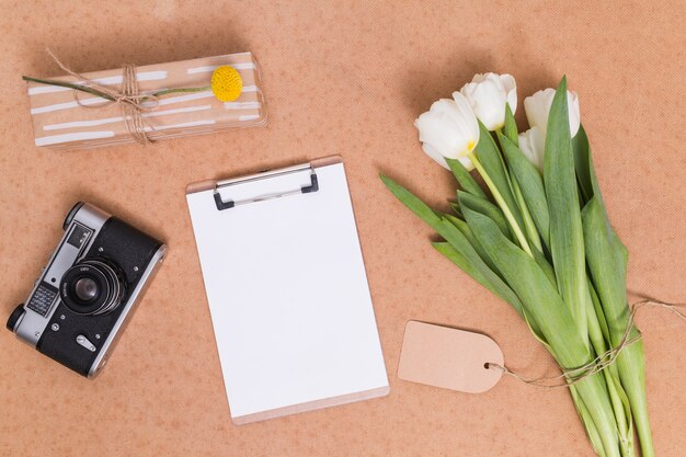 Vista elevada do grupo de flores de tulipa branca; câmera retro; caixa de presente e papel branco com prancheta na mesa